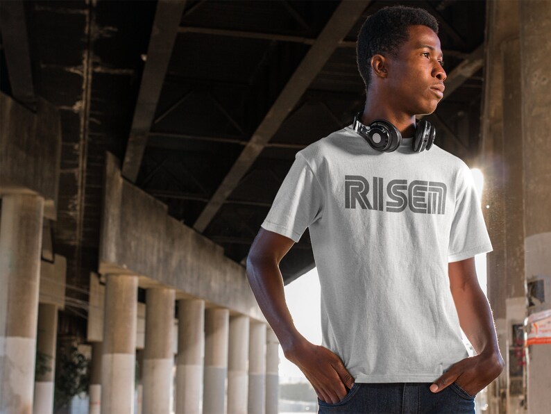 Op de afbeelding: Een wit t-shirt met het woord "RISEN" in grijs gedrukt, met een retro lettertype. Het shirt met korte mouwen wordt gedragen door een persoon met een koptelefoon om de nek. De achtergrond bevat betonnen pilaren en balken.