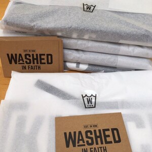 Op de afbeelding: Een stapel verpakte items verpakt in wit vloeipapier. Elke verpakking heeft een zwart-wit logo. Bruine kaarten met de tekst "WASHED IN FAITH" in zwart zijn ook zichtbaar.
