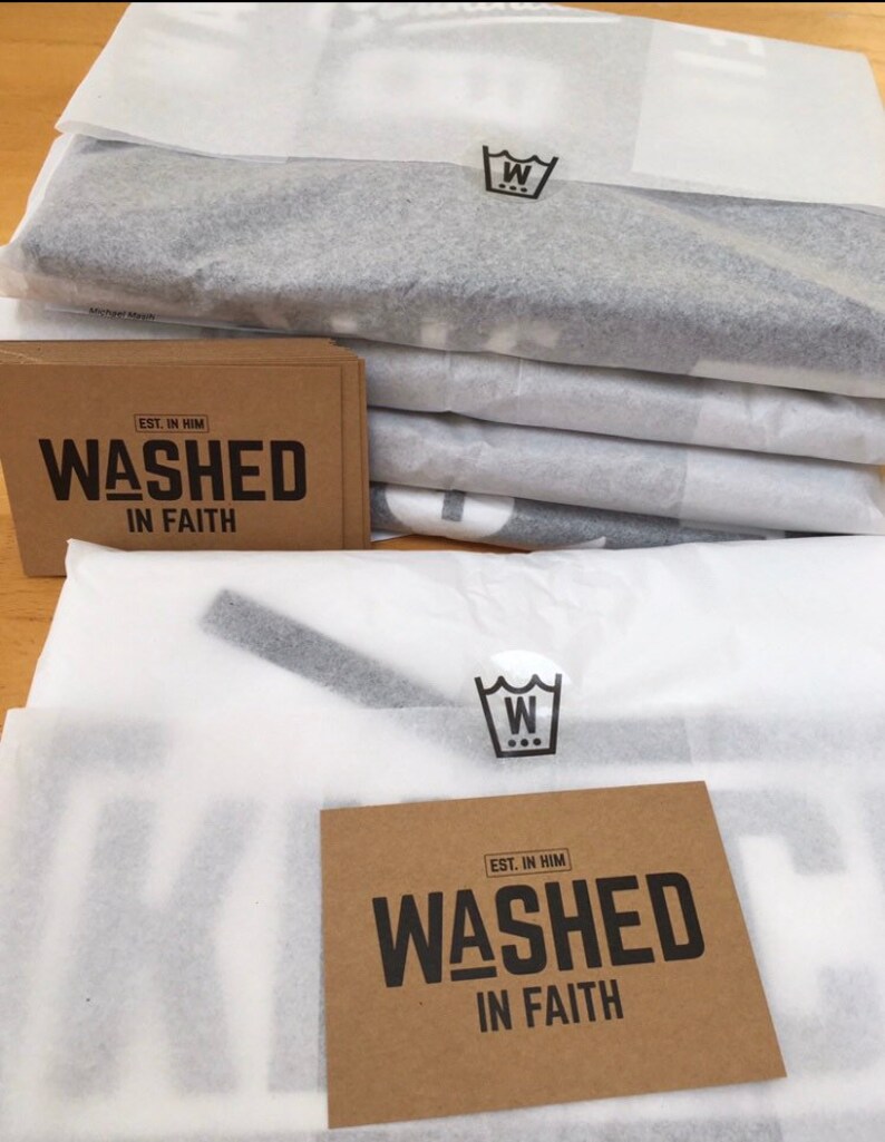 Op de afbeelding: Een stapel verpakte items verpakt in wit vloeipapier. Elke verpakking heeft een zwart-wit logo. Bruine kaarten met de tekst "WASHED IN FAITH" in zwart zijn ook zichtbaar.
