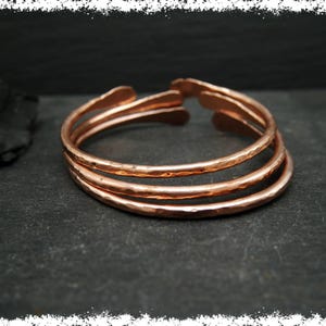 May include: Three hammered copper bangles stacked on a dark surface. The bangles are a warm copper colour and have a slightly irregular shape.