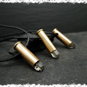 Puede incluir: Tres casquillos de bala de color dorado con cristales de cuarzo transparentes unidos, colgando de un cordón de cuero negro.