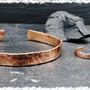Puede incluir: Dos pulseras de cobre martilladas, una más grande y otra más pequeña, sobre una superficie gris oscura. La pulsera más grande es más ancha y tiene una textura martillada más pronunciada.