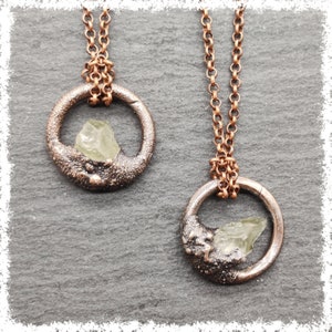 Puede incluir: Dos collares con colgantes de color cobre. Cada collar presenta un colgante circular con una superficie rugosa y texturizada y un cristal verde claro. Los colgantes cuelgan de una cadena de cobre a juego. El fondo es una superficie texturizada de color gris oscuro.