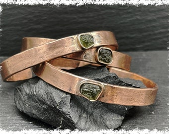 May include: Three copper bracelets with green gemstone accents. The bracelets are stacked on top of each other and are resting on a dark gray rock.