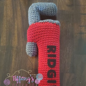 May include: A red and gray crocheted plush toy shaped like a pipe wrench. The wrench has the word "RIDGID" written on it in black.