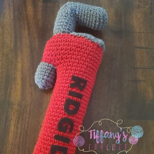 May include: A red and grey crocheted wrench with the word "RIDGID" in black lettering. The wrench is a plush toy and is handmade. The image also includes a small logo for "Tiffany's Crochet" in the bottom right corner.