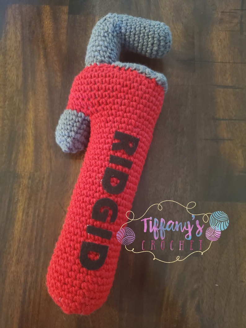 May include: A red and grey crocheted wrench with the word "RIDGID" in black lettering. The wrench is a plush toy and is handmade. The image also includes a small logo for "Tiffany's Crochet" in the bottom right corner.