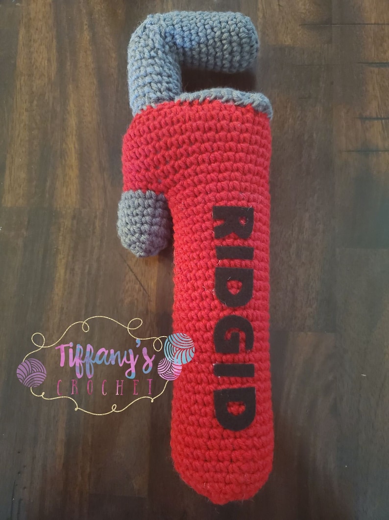 May include: A red and grey crocheted plush toy shaped like a pipe wrench. The wrench has the word "RIDGID" written on it in black.