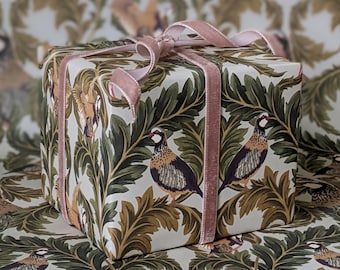 May include: A square gift box wrapped in cream-coloured paper featuring a repeating pattern of brown and black birds perched among dark green foliage. The box is tied with a soft, pale pink velvet ribbon that forms a bow on top.