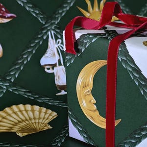 May include: A gift box wrapped in patterned paper with a red velvet ribbon. The paper features a green background with illustrations of a crescent moon, sun, and other designs. The ribbon adds a touch of elegance.
