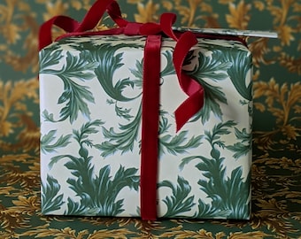 May include: A wrapped gift box with cream-colored paper featuring a green leaf pattern. The box is tied with a wide, red velvet ribbon and bow. A small tag is attached to the top of the box, suggesting a special occasion.