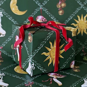 May include: A wrapped gift box with a dark green background and a repeating pattern of suns, moons, and mushrooms. The box is tied with a red velvet ribbon. The corners of the box are trimmed with gold.