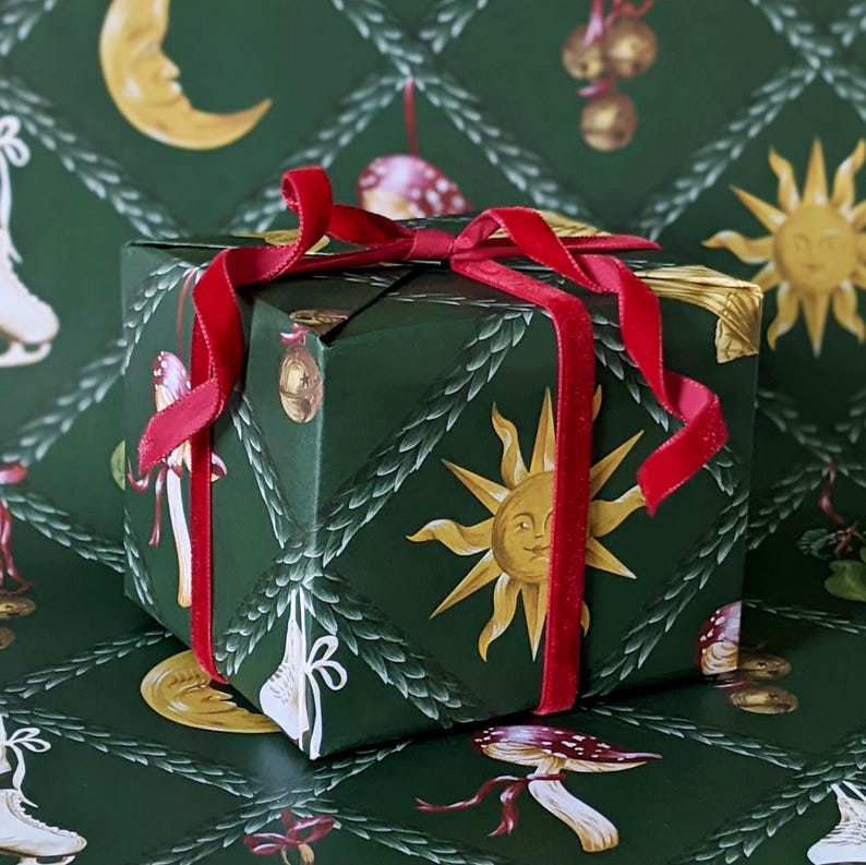 May include: A wrapped gift box with a dark green background and a repeating pattern of suns, moons, and mushrooms. The box is tied with a red velvet ribbon. The corners of the box are trimmed with gold.