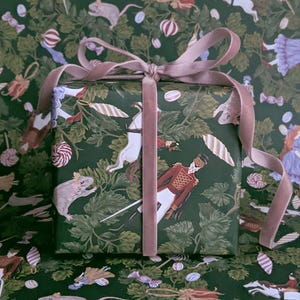 May include: A square gift box wrapped in dark green paper featuring a repeating pattern of nutcracker figures, white horses, and sweets. The box is tied with a dusty pink velvet ribbon that is knotted in a bow on the top centre of the package.