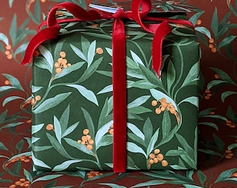 May include: A wrapped gift box with a dark green botanical print featuring leaves and berries. The box is tied with a wide, deep red velvet ribbon. A matching gift tag is attached. The background is a similar pattern on a brown surface.