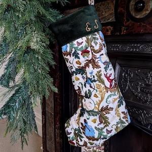 May include: A Christmas stocking featuring a dark green velvet cuff embroidered with the gold letters E and S. The body of the stocking is made of white fabric decorated with a colourful pattern of foliage, pears, and small figures in various poses.