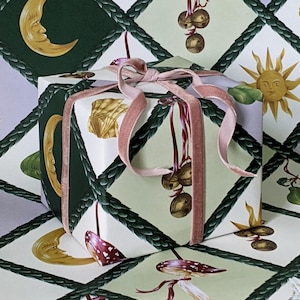 May include: A square gift box wrapped in patterned paper with a light pink velvet ribbon. The paper design includes moons, suns, and mushrooms in green, gold, and white. The ribbon is tied in a bow around the box.