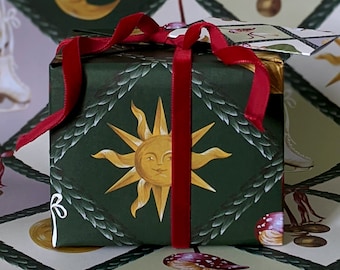 May include: A wrapped gift box with a dark green background featuring a smiling sun design in gold. The box is tied with a red velvet ribbon. The wrapping paper has a repeating pattern of moons, suns, and mushrooms.