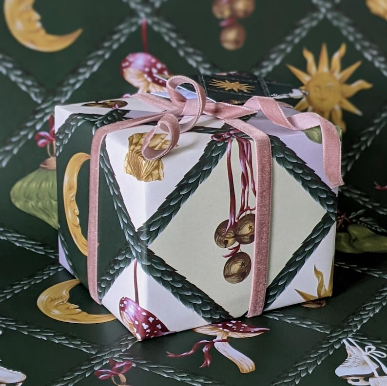 May include: A gift box wrapped in patterned paper with moons, suns, and mushrooms. The box is tied with a pink velvet ribbon. The background is a dark green with a repeating pattern. The overall design has a whimsical, fairytale aesthetic.