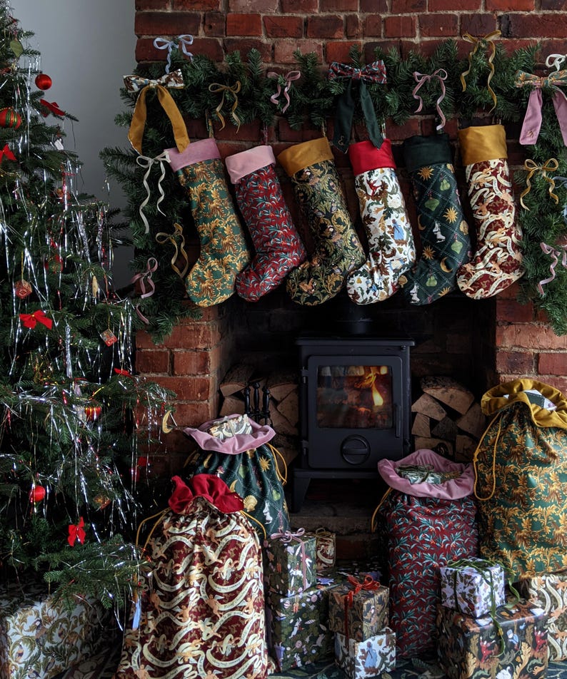 May include: A Christmas display with a decorated tree, stockings, and gifts. Six patterned stockings hang above a fireplace with a lit fire. Gift bags and wrapped presents are arranged in front of the fireplace, creating a festive holiday scene.