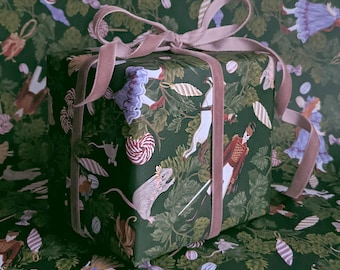 May include: A square gift box wrapped in dark green paper featuring a repeating pattern of Nutcracker-themed characters, mice, and candy, tied with a soft, light pink velvet ribbon bow on top.