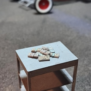 Może przedstawiać: Miniaturowy szary stolik z dwoma półkami, na którym leżą stosy zabawkowych pieniędzy. Rozmyte tło przedstawia czerwono-biały samochodzik. Scena sugeruje miniaturową ekspozycję, prawdopodobnie związaną z bogactwem.