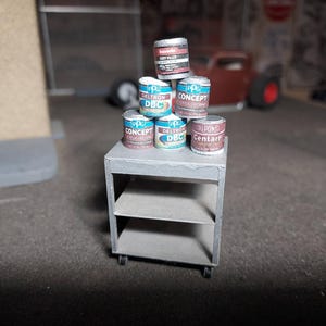Puede incluir: Una estantería metálica gris en miniatura con varias latas de pintura pequeñas apiladas encima. Las latas están etiquetadas con varias marcas y colores, incluyendo "Deltron DBC" y "Concept". El fondo muestra una escena callejera borrosa.