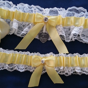 May include: Two yellow satin garters with white lace trim. The top garter has a silver heart charm with the text "Albert & Christine 2019".