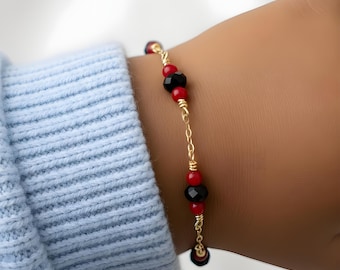 May include: A delicate gold-tone chain bracelet featuring alternating small red beads and larger faceted black beads. The bracelet is constructed with wire-wrapped links connecting the beads, creating a repeating pattern of red and black accents.