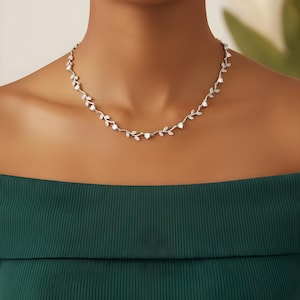 Puede incluir: Un collar plateado con un delicado diseño de vid. El collar presenta pequeñas piedras preciosas transparentes y detalles en forma de hoja. El collar lo lleva una persona que lleva una blusa verde oscuro con los hombros descubiertos.