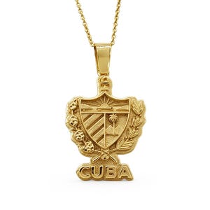 May include: A gold-colored pendant necklace featuring a shield design with the word "CUBA" at the bottom. The shield has a sunburst, palm tree, and other details. The necklace chain is also gold.