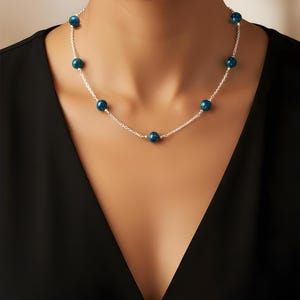 May include: A silver necklace featuring a delicate chain and a series of round, dark teal beads. The beads have a speckled appearance, and the necklace is displayed against a black top.
