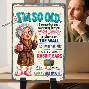 May include: A rectangular slate plaque with a rough edge featuring a cartoon of an elderly person in a brown robe and bunny slippers holding a coffee mug. The text reads: I'M SO OLD, I remember one bathroom for the whole family, a phone on THE WALL, no internet, & a TV with RABBIT EARS & just 3 channels AND WE STILL MADE IT.