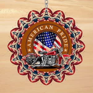 May include: Round ornament with a red motorcycle graphic, the American flag, and the words "AMERICAN PRIDE." The ornament is surrounded by a decorative floral design in red, blue, and brown, and is suspended by a silver chain.