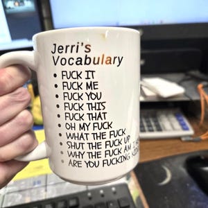 May include: A white ceramic mug featuring the black printed text Jerri's Vocabulary followed by a list of phrases including FUCK IT, FUCK ME, FUCK YOU, FUCK THIS, FUCK THAT, OH MY FUCK, WHAT THE FUCK, SHUT THE FUCK UP, and WHY THE FUCK AM I HERE.
