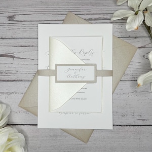 May include: A white wedding invitation with a beige envelope. The invitation has a satin ribbon with a small tag that says "Jennifer & Anthony".