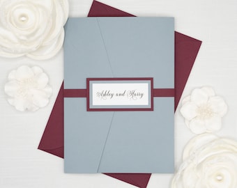 May include: A gray wedding invitation with a burgundy envelope. The invitation has a burgundy ribbon and a white label with the text "Ashley and Harry" in black script.