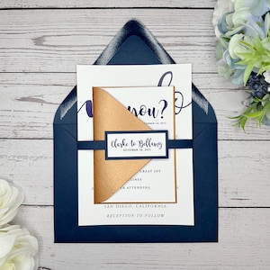 May include: Navy blue wedding invitation envelope with a white card inside. The card has a gold and brown inner envelope with a white label that says "Clarke to Bellamy" and the date "October 18, 2015".