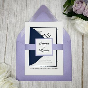 May include: A lavender wedding invitation with a navy blue inner flap. The invitation features the text "Olivia & Kevin" in a script font. The invitation is on a white wooden background.