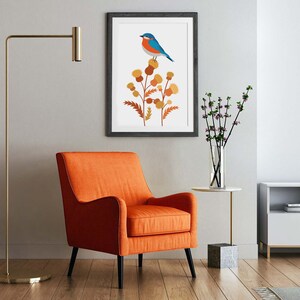 May include: An orange armchair with black legs sits on a wooden floor. A framed print of a blue bird perched on a plant hangs on the wall. A gold floor lamp and a small side table with a vase of flowers complete the room.