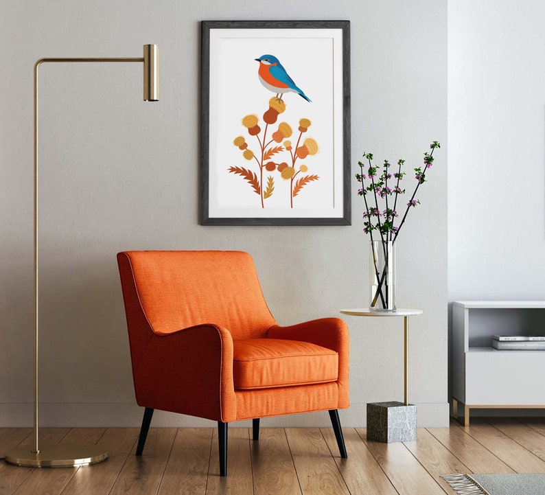 May include: An orange armchair with black legs sits on a wooden floor. A framed print of a blue bird perched on a plant hangs on the wall. A gold floor lamp and a small side table with a vase of flowers complete the room.