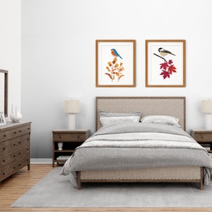 May include: Two framed art prints featuring birds. One print shows a blue bird perched on orange and yellow flowers. The other print depicts a black and white bird on a branch with red leaves. The frames are brown, and the prints are displayed above a bed.