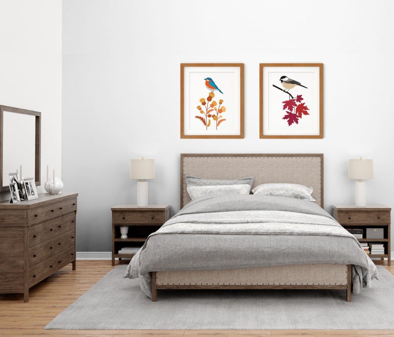 May include: Two framed art prints featuring birds. One print shows a blue bird perched on orange and yellow flowers. The other print depicts a black and white bird on a branch with red leaves. The frames are brown, and the prints are displayed above a bed.