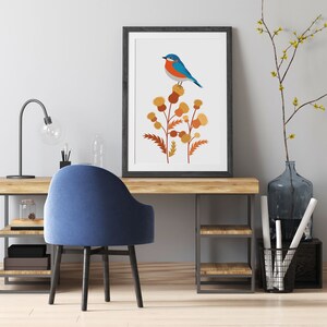 May include: A framed art print featuring a blue and orange bird perched on a cluster of stylized brown and yellow flowers. The artwork is displayed on a wooden desk with a blue chair, a black desk lamp, and decorative items.
