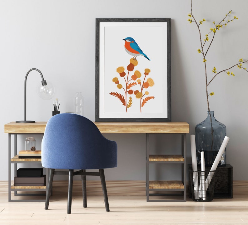 May include: A framed art print featuring a blue and orange bird perched on a cluster of stylized brown and yellow flowers. The artwork is displayed on a wooden desk with a blue chair, a black desk lamp, and decorative items.