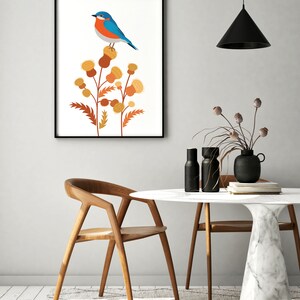 May include: A framed art print featuring a blue and orange bird perched on stylized yellow and brown thistle flowers. The artwork is displayed in a modern interior setting with a wooden chair and a round table with a marble top.