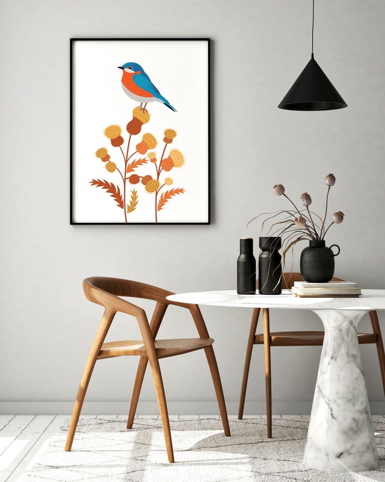 May include: A framed art print featuring a blue and orange bird perched on stylized yellow and brown thistle flowers. The artwork is displayed in a modern interior setting with a wooden chair and a round table with a marble top.