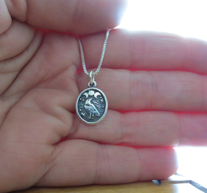 May include: A silver necklace with a round pendant featuring a raven with three crescent moons and stars. The pendant is on a silver chain.