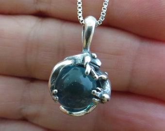 Puede incluir: Collar con colgante de plata con una pequeña rana sosteniendo una esfera de vidrio azul.