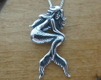 May include: A silver mermaid pendant necklace with a detailed design. The mermaid is sitting with her tail curled behind her. The pendant is on a silver chain.